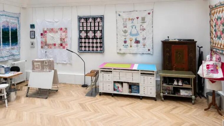 Atelier de couture sous verrière : table de coupe Auboi au centre de la pièce, parquet à chevrons et panneaux d’ouvrages au mur