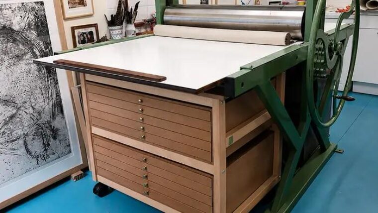 Presse taille-douce en fonte verte accolée à un meuble à plans Auboi, estampes encadrées au mur de l’atelier