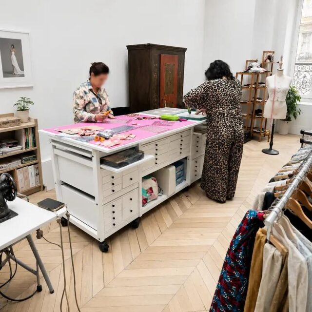 Atelier de couture sous verrière : table de coupe Auboi au centre, deux couturières au travail, parquet à chevrons