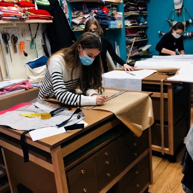 Association La Refile : une couturière trace un patron sur une table atelier Auboi, tiroirs de mercerie au-dessous